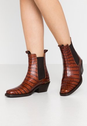 Botas de tobillo de charol en color marrón con estampado de cocodrilo y paneles laterales elásticos, colocadas sobre una superficie blanca brillante. Piernas parcialmente visibles, mostrando las botas de perfil.