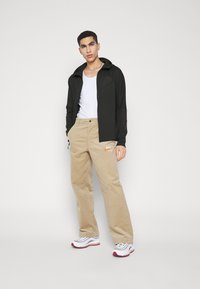 Czarna bluza z zamkiem błyskawicznym, beżowe spodnie z cordurowego materiału z logo Nike. Jasny podkoszulek. Sneakersy z czerwonymi akcentami. Gładkie materiały, luźny krój.