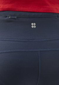 Navy Leggings mit einer glatten Textur, ausgestattet mit einer kleinen Reißverschlusstasche und einem silbernen Logo. Die Nähte verleihen dem Design zusätzliche Akzente.