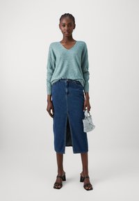 Τιρκουάζ πλεκτό πουλόβερ με λαιμόκοψη σε V, συνδυασμένο με μεσαίου μήκους τζιν φούστα με μπροστινή σχίσιμο. Κρατάει μια μικρή ανοιχτό μπλε τσάντα. Μαύρα παπούτσια με τακούνι.