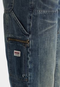 Denim-Jeans-Tasche mit messingfarbendem Reißverschluss, Nieten und einem weißen Guess Originals Stoffetikett mit rotem und schwarzem Text.