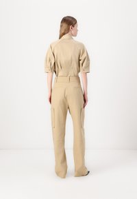 Beige utility jumpsuit with short sleeves, button details on sleeves, and cargo pockets on pants. Textured fabric, relaxed fit.