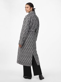 Cappotto lungo in tessuto a scacchi in bianco e nero, con cintura, spacchi laterali e un ampio colletto. Indossato sopra pantaloni neri a gamba larga e scarpe.