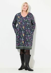 Långärmad klänning med V-ringning i mörk tyg som har ett färgglatt paisleymönster och fickor, matchad med höga svarta cowboyboots.