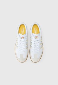 Baskets blanches avec des rayures jaunes et grises, bout en daim texturé et semelle en gomme. Présente le logo Adidas et une marque sur la languette.