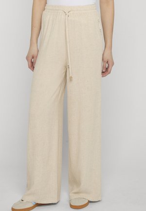 Beige Leinenhose mit weitem Bein, elastischem Bund und Kordelzug, kombiniert mit weißem Oberteil und hellbeigen Schuhen mit braunen Sohlen.