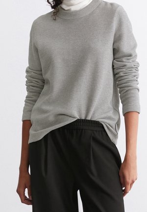 Persoon draagt een gestructureerde lichtgrijze trui met lange mouwen over een witte coltrui en losse zwarte broek met elastische tailleband.