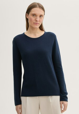 Marineblå langærmet sweater med rund halsudskæring og tekstureret stof, med lige snit og afslappet pasform, parret med lyse bukser.