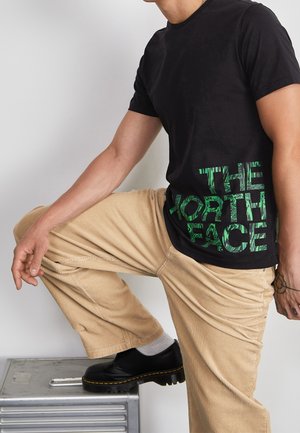 Camiseta negra de manga corta con texto gráfico verde "THE NORTH FACE." Combinada con pantalones beige de pana y calzado negro.