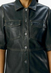 Zwarte leren shirtjacket met borstzakken, zilveren knopen en contrasterende stiksels. Korte mouwen met een gestructureerd kraagontwerp.