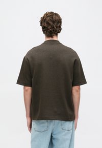 Kortermet, mørke olivengrønn skjorte med rund hals og avslappet passform, laget av teksturert stoff. Brukt med lys blå denimjeans.