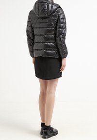 Black puffer jacket with a shiny finish, hood, and horizontal quilting; worn over a black skirt, viewed from the back, with black shoes.