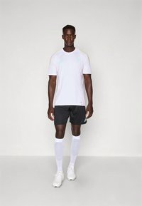 Balts īss piedurkņu sporta krekls ar mīksti zaļiem akcentiem, kopā ar melniem šortiem un baltiem ceļgalu augstiem zeķēm, stāvot uz vienkārša fona.