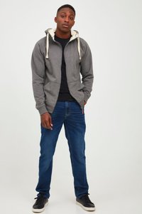 Felpa grigia con zip e cordini crema, t-shirt nera e jeans blu. Il modello si trova con le mani in tasca, indossando sneakers nere.