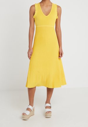 Femme portant une robe midi plissée jaune sans manches avec des détails à volants et des sandales compensées blanches sur un fond uni.