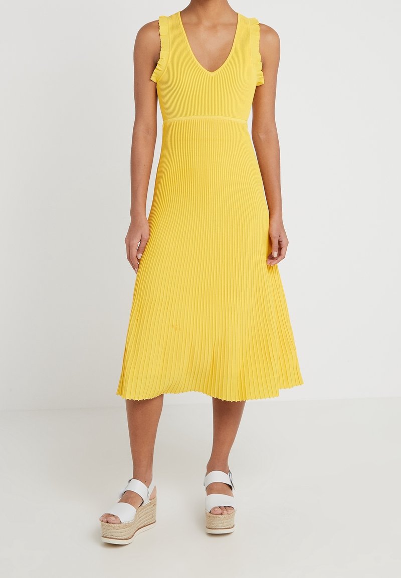 Femme portant une robe midi plissée jaune sans manches avec des détails à volants et des sandales compensées blanches sur un fond uni.