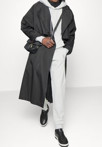Largo abrigo negro tipo trench con un diseño a medida, superpuesto a una sudadera gris clara y pantalones de chándal. Zapatillas negras con acentos texturizados.