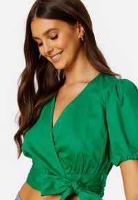 Top corto verde a portafoglio con maniche a palloncino, realizzato in un tessuto morbido. Caratterizzato da una profonda scollatura a V e un dettaglio annodato in vita.