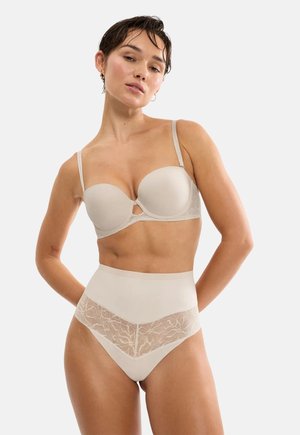 Kvinde iført beige balconette-bh og højtaljet trusse med blonde, stående mod en ensfarvet lys baggrund.