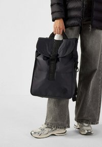 Zaino in nylon nero con chiusura a patta, tracolla regolabile, tasca frontale e dettagli del manico con marchio, completato da una texture liscia.