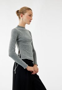 Grijze geribde trui met lange mouwen en een opstaande kraag. Heeft zijslits met verstelbare strikken en een cropped zoom. Draagt in combinatie met zwarte broek.