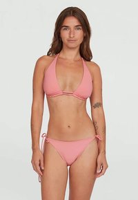 Γυναίκα με μακριά καστανά μαλλιά, φορώντας ένα ανοιχτό ροζ μπικίνι με λαιμόκοψη και παντελόνι με κορδόνια στο πλάι, που στέκεται μπροστά σε μια απλή λευκή οθόνη.