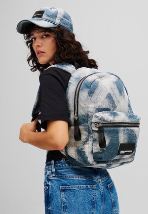 Zaino in denim con un motivo astratto blu e bianco, caratterizzato da dettagli neri zippati e una tasca frontale. Completato da un cappellino abbinato.