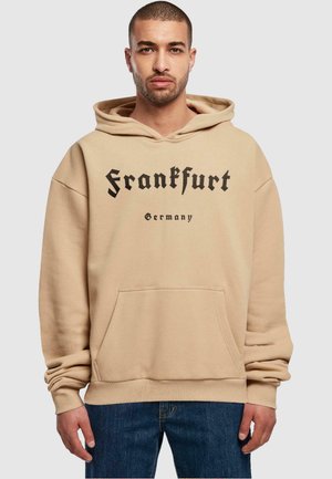 Beige hettegenser med kengurulomme, med svart tekst "Frankfurt" og "Tyskland" trykket i en fet font. Kvalitet i bomullsblanding, avslappet passform.