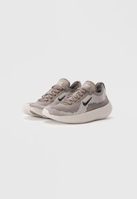 Lysbrune og grå strikkesneakers med et tekstureret mønster, der har sorte logo accenter og en hvid gummisål. Snørebåndsdesign, lav profil.