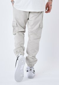 Pantaloni cargo beige con polsini elastici, tasche laterali e una texture liscia, abbinati a sneakers bianche e nere.