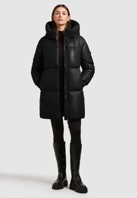 Vrouw die een zwarte gewatteerde jas met capuchon draagt, zwarte legging en zwarte laarzen tot aan de knie, staand tegen een eenvoudige lichte achtergrond.