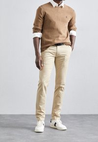 Pull en tricot marron, chemise blanche à col en dessous, chinos beige et baskets blanches avec des accents noirs. Design simple et silhouette ajustée.