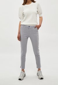 Žena v bílém textured topu s krátkým rukávem, tmavě modrých a bílých vertikálně pruhovaných slim kalhotách a světle šedých teniskách, stojící s jednou rukou v kapse.