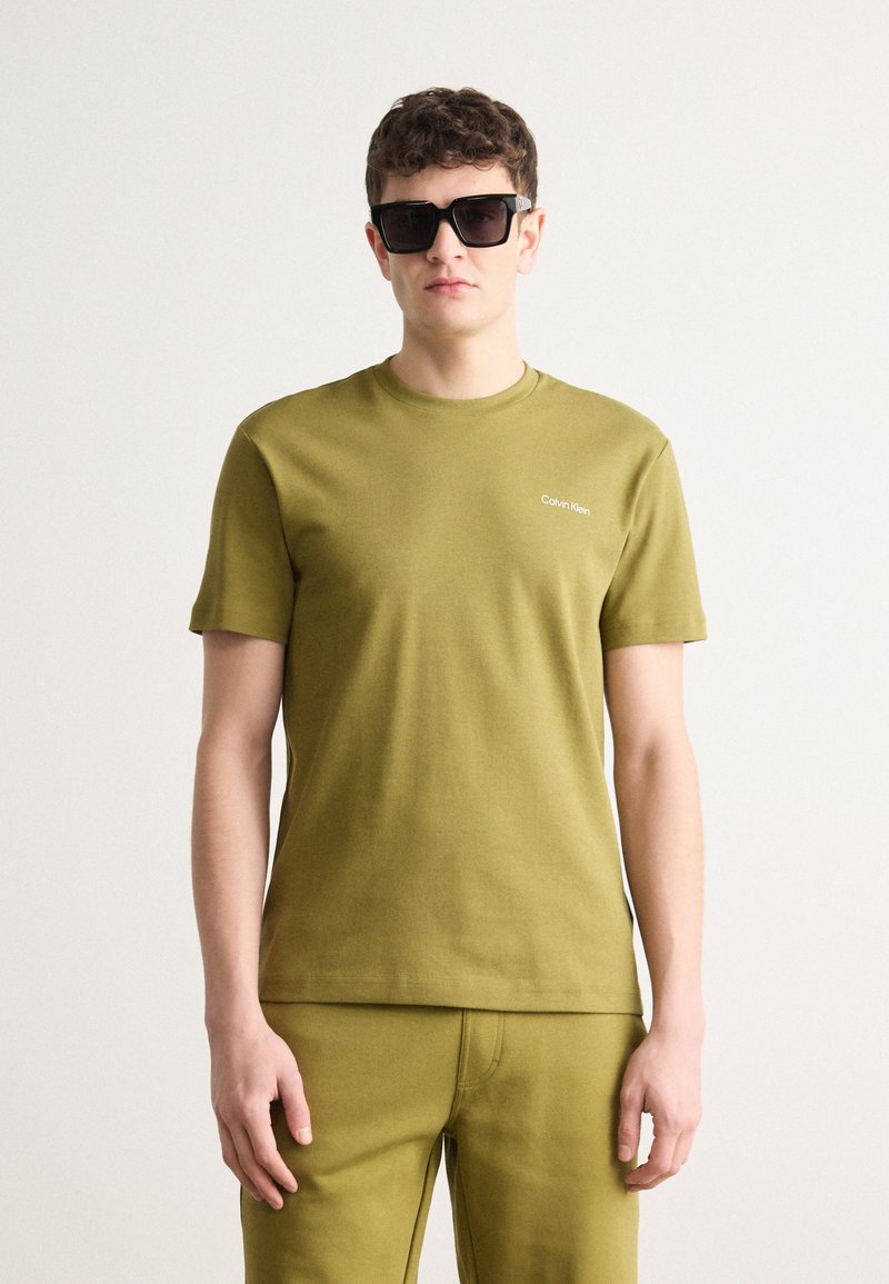 Tricou din bumbac de culoare verde măslin, cu guler rotund și mâneci scurte, având un mic logo alb "Calvin Klein" pe piept.