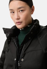 Zwarte gewatteerde jas met een hoge kraag, voorzien van een ritssluiting en metalen knopen. Draagt over een groene coltrui met witte patroonaccenten.