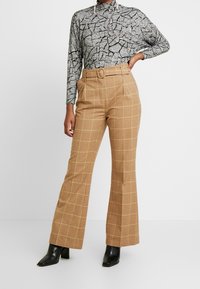 Top gris de manga larga con un patrón de grietas, combinado con pantalones de cuadros marrones de talle alto con líneas amarillas y piernas acampanadas. Botines negros.