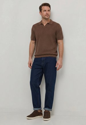 Mann trägt ein braunes kurzärmeliges Strick-Poloshirt, dunkelblaue hochgekrempelte Jeans und braune Sneakers und steht vor einer hellgrauen Wand.