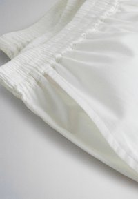 Close-up van witte stoffen shorts met een elastische tailleband en een zichtbare zijzak op een witte achtergrond.