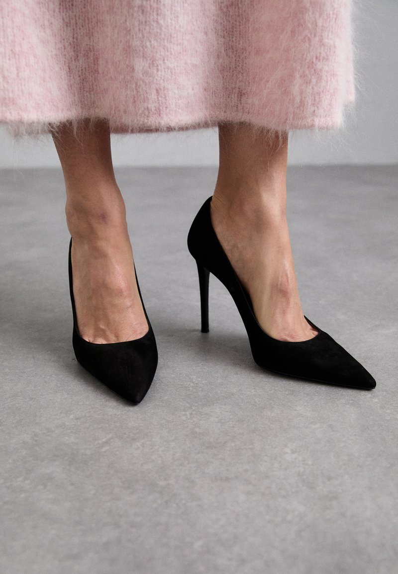 Zwarte suède hoge hakken met puntige neuzen en stiletto-hakken. Draagt met een roze fluffy rok, de focus ligt op het ontwerp en de textuur van de schoenen.