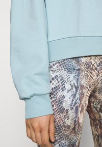 Felpa azzurro chiaro con polsini a coste abbinata a leggings marroni e beige con motivo a serpente. Tessuto liscio con design aderente.