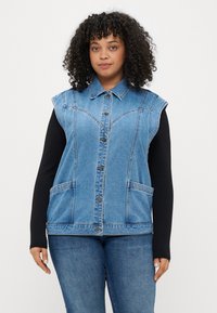 Jeans brezrokavnik svetlo modre barve z gumbnim zapiranjem, dvema sprednjima žepoma in šivnimi detajli. Nošen preko črne majice z dolgimi rokavi.