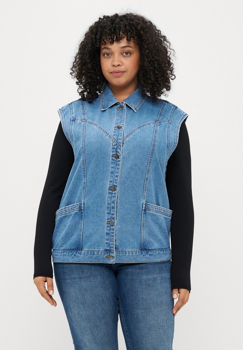 Jeans brezrokavnik svetlo modre barve z gumbnim zapiranjem, dvema sprednjima žepoma in šivnimi detajli. Nošen preko črne majice z dolgimi rokavi.