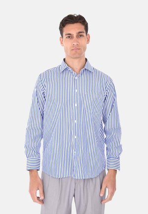 Uomo che indossa una camicia a maniche lunghe con righe verticali blu e bianche e pantaloni grigi chiari, in piedi davanti a uno sfondo semplice.