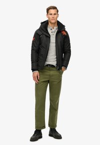 Czarna kurtka puchowa z zamkami, kapturem i pomarańczowymi akcentami logo, noszona na jasnoszarym henleyu, zestawiona z zielonymi spodniami i czarnymi butami.