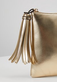 Kultainen metallinhohtoinen clutch-laukku, jossa sileä pinta, vetoketjukiinnitys ja koristeellinen hapsutettu tupsu lisäilmettä varten.