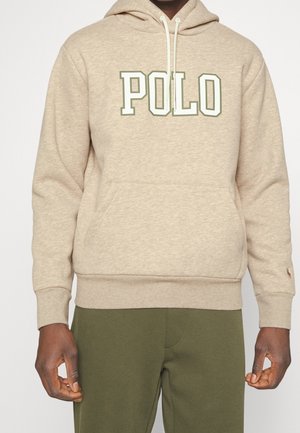 Beige hættetrøje med "POLO" tekst i grønne og hvide blokbogstaver, båret med olivengrønne bukser, med forlommekangorulomme og snøre på hætten.