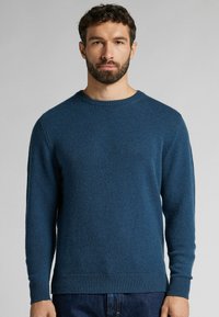 Blauwe gebreide trui met een ronde halslijn, lange mouwen en ribgebreide manchetten en onderrand. Zachte textuur, casual pasvorm. Draagt met donkere spijkerbroek.