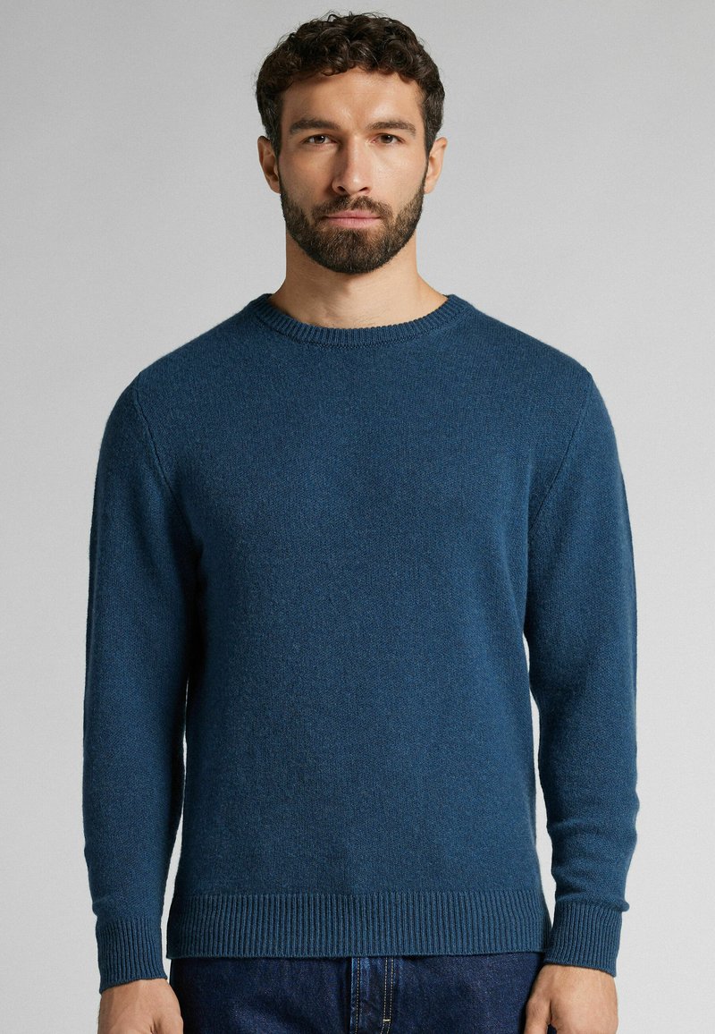 Blauwe gebreide trui met een ronde halslijn, lange mouwen en ribgebreide manchetten en onderrand. Zachte textuur, casual pasvorm. Draagt met donkere spijkerbroek.