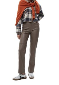 Braune Kunstlederhose mit hoher Taille und schmal zulaufendem Bein, kombiniert mit einem karierten Button-Down-Hemd und einem über die Schultern gelegten orangefarbenen Pullover.