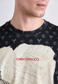 Sort sweatshirt med et trykt kortdesign i beige, med teksten "CARLO COLUCCI" i rødt og et subtilt logomønster over det hele.